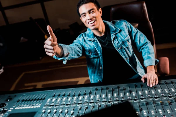 Smiling mixed race sound producer showing thumb up while standing near mixing console in recording studio — Stock Photo