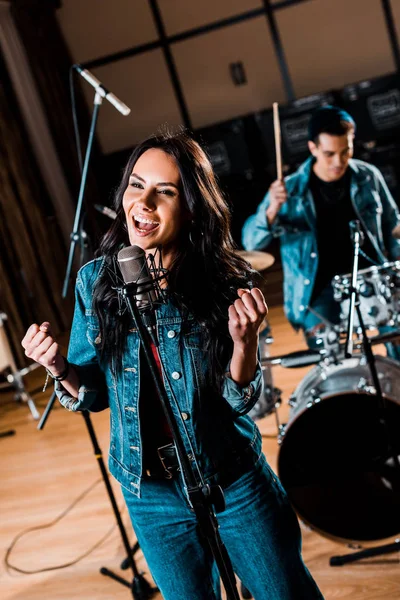 Messa a fuoco selettiva di bella donna emotiva che canta mentre musicista di razza mista suona la batteria in studio di registrazione — Foto stock