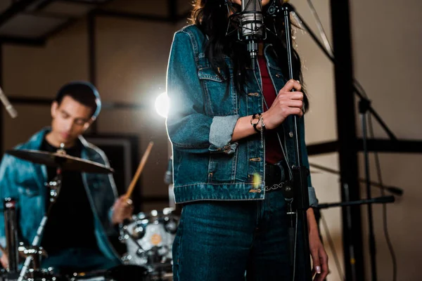 Selective focus of woman singing in recording studio while mixed race musician playing drums — Stock Photo
