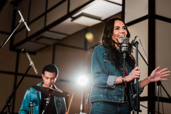 Selective focus of excited woman singing in recording studio while handsome mixed race musician playing drums — Stock Photo