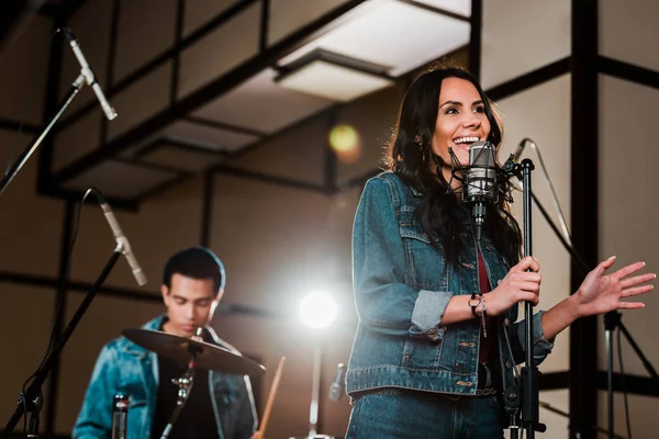 Enfoque selectivo de la mujer emocionada cantando en el estudio de grabación mientras guapo músico de raza mixta tocando la batería - foto de stock