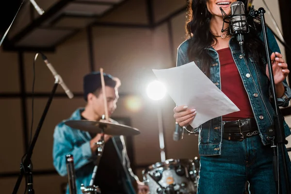 Partial view of smiling woman singing in recording studio while mixed race musician playing drums — Stock Photo