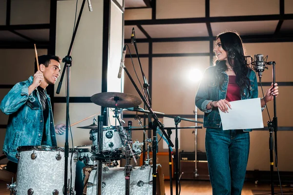 Bonito músico de raça mista na bateria e atraente cantor perto de microfone em estúdio de gravação — Fotografia de Stock