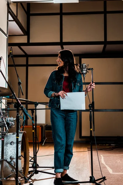 Schöne Sängerin steht neben Mikrofon und hält Papier mit Text im Tonstudio — Stockfoto