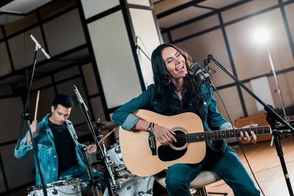 Selective focus of beautiful inspired musician playing guitar and singing while mixed race man playing drums in recording studio — Stock Photo