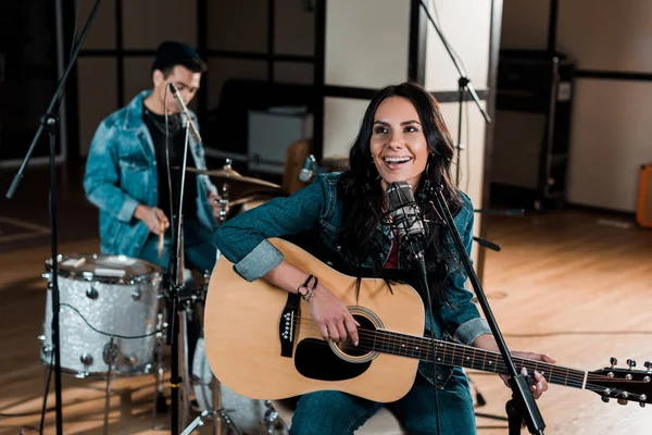 Selective focus of beautiful woman playing guitar and singing while mixed race musician playing drums — Stock Photo