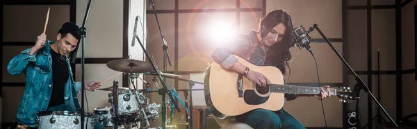 Panoramic shot of attractive woman playing guitar while mixed race musician playing drums in recording studio — Stock Photo