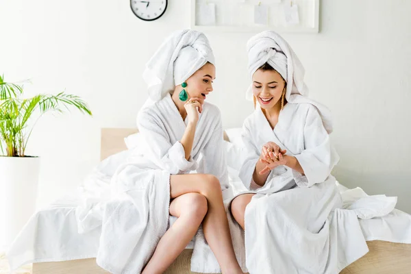 Chicas elegantes en pendientes, albornoces y con toallas en la cabeza hablando mientras está sentado en la cama - foto de stock