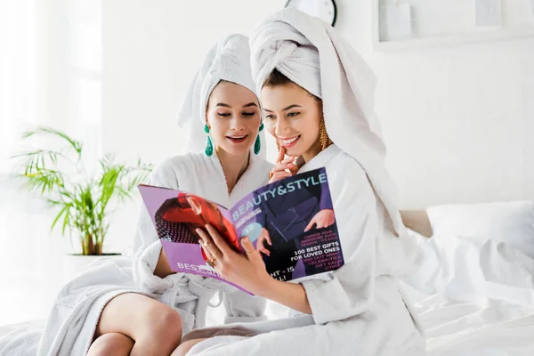 Chicas elegantes en pendientes, albornoces y con toallas en las cabezas de estilo de lectura y revista de belleza mientras está sentado en la cama - foto de stock
