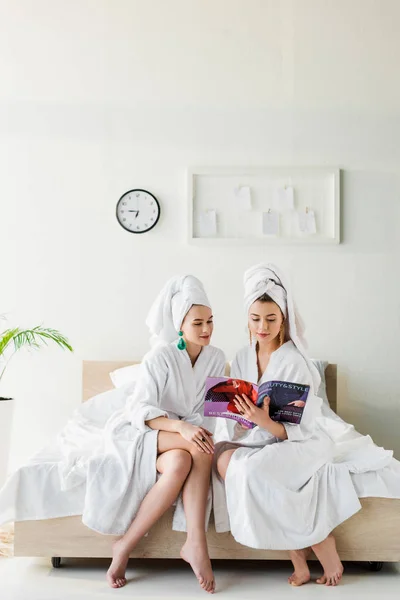Femmes élégantes dans des boucles d'oreilles, peignoirs et avec des serviettes sur les têtes style de lecture et magazine de beauté tout en étant assis sur le lit — Photo de stock