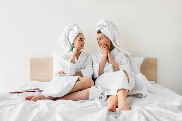 Meninas elegantes em brincos, roupões de banho e com toalhas na cabeça sentados na cama e olhando um para o outro — Fotografia de Stock