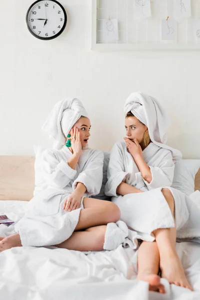 Meninas elegantes chocadas em brincos, roupões de banho e com toalhas na cabeça sentados na cama e olhando um para o outro — Fotografia de Stock