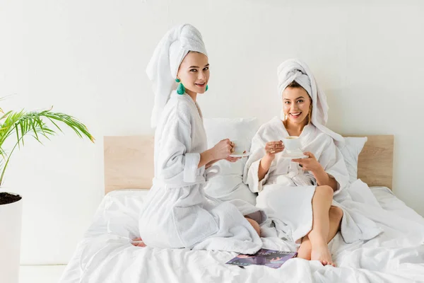 Donne eleganti in accappatoi, orecchini e con asciugamani sulla testa guardando la fotocamera e tenendo tazze di caffè e piattini a letto — Foto stock