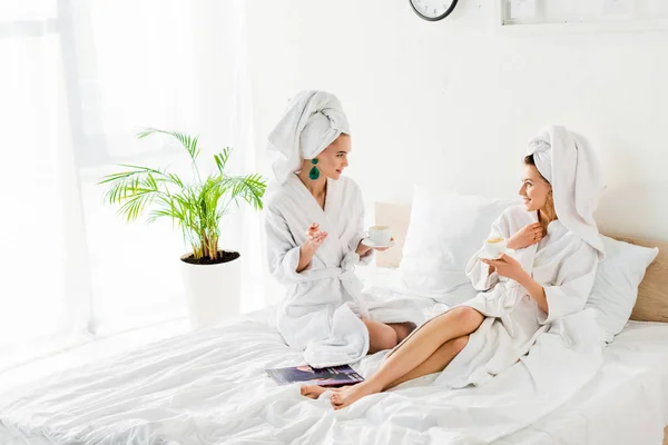 Élégantes femmes en peignoirs, boucles d'oreilles et avec des serviettes sur les têtes tenant des tasses à café et des soucoupes tout en parlant au lit — Photo de stock