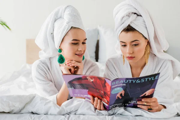 Mulheres elegantes em roupões de banho, brincos e com toalhas na cabeça ler revista enquanto deitado na cama — Fotografia de Stock