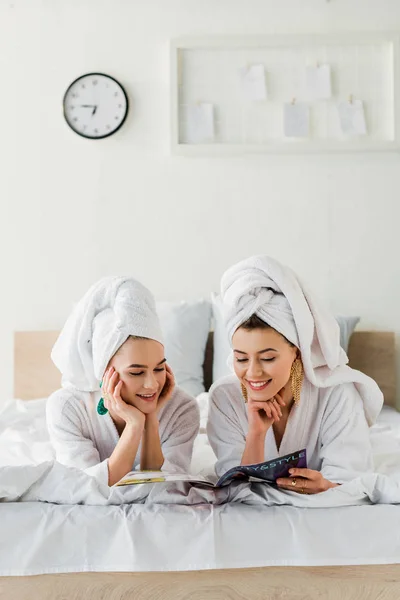 Happy stylish women in bathrobes, earrings and with towels on heads reading magazine while lying in bed — Stock Photo