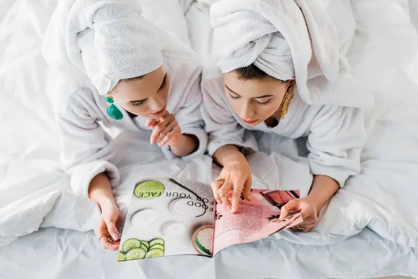 Vista aérea de las mujeres con estilo en batas de baño, pendientes y con toallas en la cabeza que leen revista juntos mientras están acostados en la cama - foto de stock