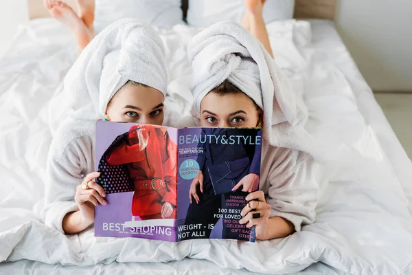 Overhead view of barefoot women in bathrobes and with towels on heads hiding behind magazine together while lying in bed — Stock Photo