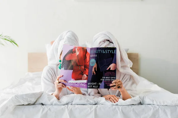 Donne in accappatoi e gioielli, con asciugamani sulla testa nascosti dietro la rivista insieme mentre si trovano a letto — Foto stock