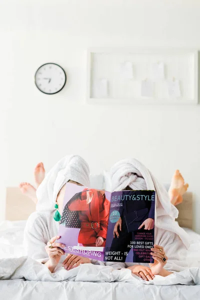Barefoot women in bathrobes and jewelry, with towels on heads hiding behind magazine together while lying in bed — Stock Photo