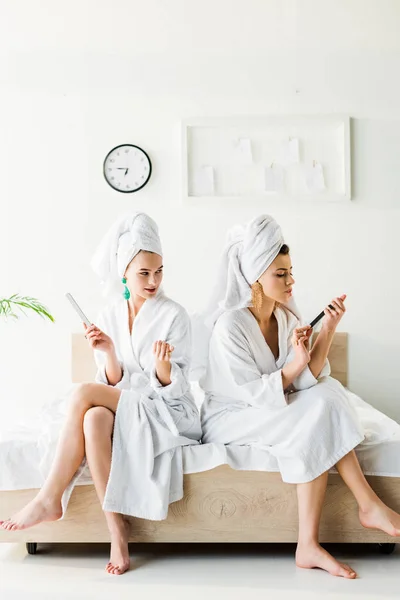 Femmes pieds nus en peignoirs et bijoux, avec des serviettes sur les têtes à l'aide de limes à ongles tout en étant assis sur le lit — Photo de stock