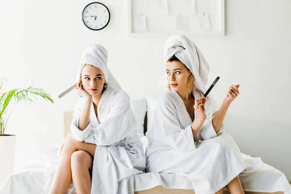 Mujeres elegantes en albornoces y joyas, con toallas en las cabezas sentadas en la cama con limas de uñas — Stock Photo