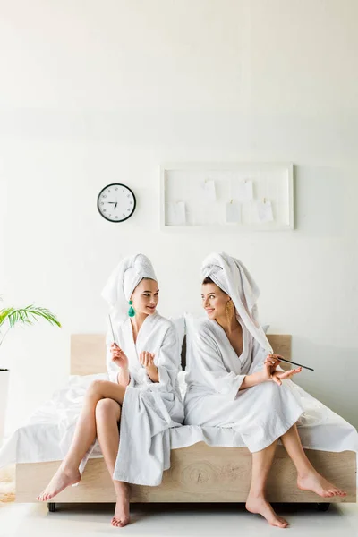 Elegantes mujeres sonrientes en albornoces y joyas, con toallas en las cabezas sentadas en la cama con limas de uñas - foto de stock