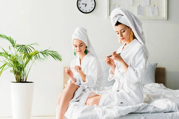 Stylish women in bathrobes and jewelry, with towels on heads sitting on bed and filing nails near green plant — Stock Photo