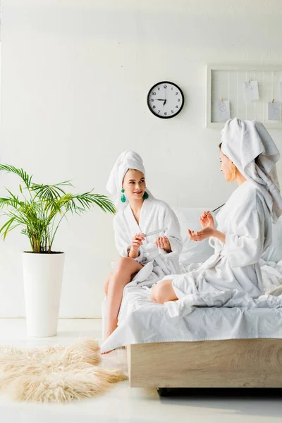 Стильные женщины в халатах и ювелирных изделиях, с полотенцами на головах, сидя на кровати, разговаривая и подавая ногти рядом с зеленым растением и мехом на полу — стоковое фото