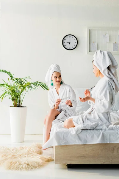 Femmes élégantes dans les peignoirs et les bijoux, avec des serviettes sur les têtes assises sur le lit, parler et limer les ongles près de plantes vertes et de fourrure sur le sol — Photo de stock