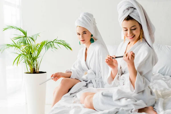 Femmes élégantes en peignoirs et bijoux, avec des serviettes sur les têtes assises sur le lit avec des limes à ongles près de la plante verte — Photo de stock