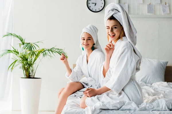 Glücklich zwinkernde stilvolle Frauen in Bademänteln und Schmuck, mit Handtüchern auf Köpfen, die mit Nagelfeilen in der Nähe grüner Pflanzen auf dem Bett sitzen und in die Kamera schauen — Stockfoto