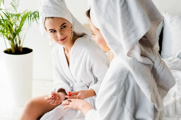 Stylish woman in bathrobe and jewelry, with towel on head sitting on bed with nail polish and looking at friend — Stock Photo