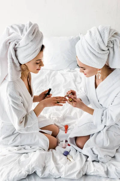 Femmes élégantes souriantes en peignoirs et bijoux, avec des serviettes sur les têtes assises sur le lit et des ongles de polissage — Photo de stock