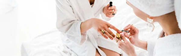Panoramic shot of women in bathrobes and jewelry sitting on bed and polishing nails — Stock Photo