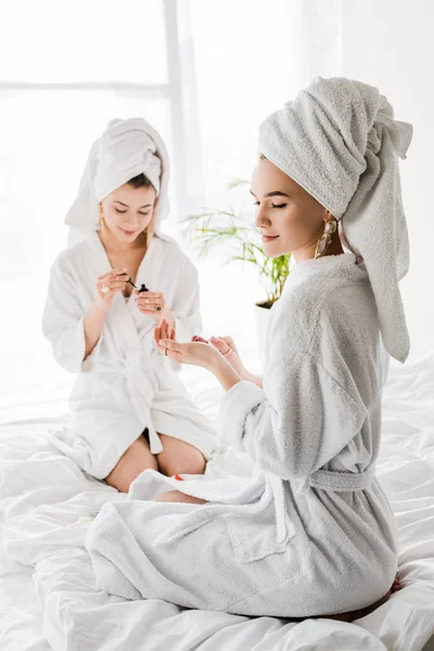 Улыбающиеся женщины в халатах, полотенцах и украшениях сидят на кровати и полируют ногти вместе — стоковое фото