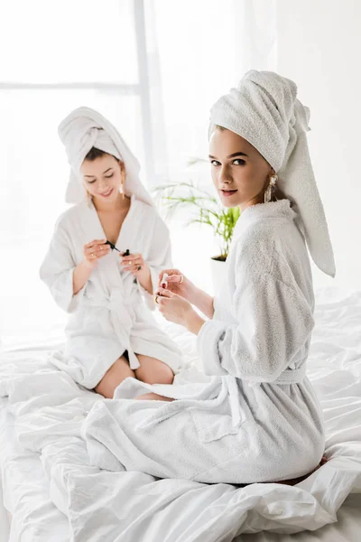 Stylish women in bathrobes, towels and jewelry sitting on bed and polishing nails together — Stock Photo