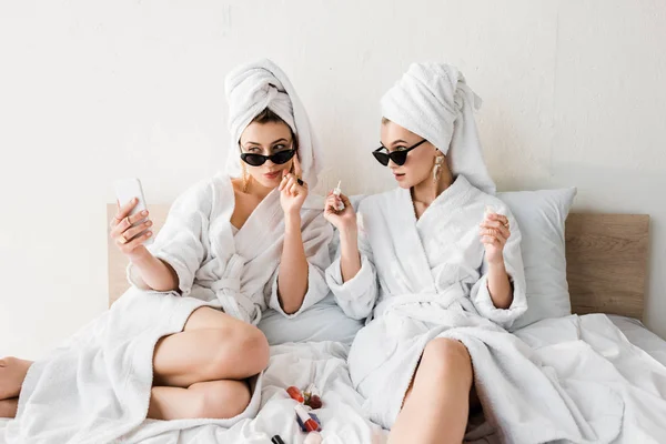 Femmes élégantes en peignoirs et lunettes de soleil, serviettes et bijoux faire pédicure et prendre selfie au lit — Photo de stock