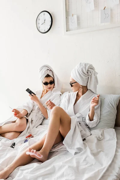 Mujeres elegantes en albornoces y gafas de sol, toallas y joyas tumbadas en la cama, haciendo pedicura y fumar - foto de stock