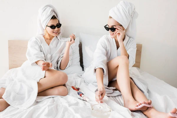 Mujeres con estilo feliz en albornoces y gafas de sol, toallas y joyas tumbadas en la cama, haciendo pedicura y fumar cerca del cenicero - foto de stock