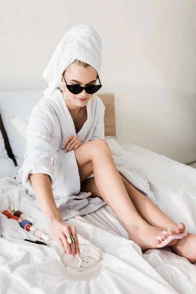 Mujer con estilo en albornoz y gafas de sol, toalla y joyas tumbado en la cama con cenicero y haciendo pedicura - foto de stock