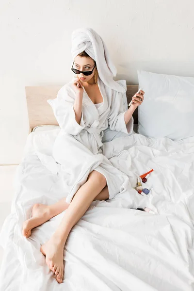 Mujer con estilo en albornoz y gafas de sol, toalla y joyas tumbado en la cama y el uso de teléfono inteligente - foto de stock