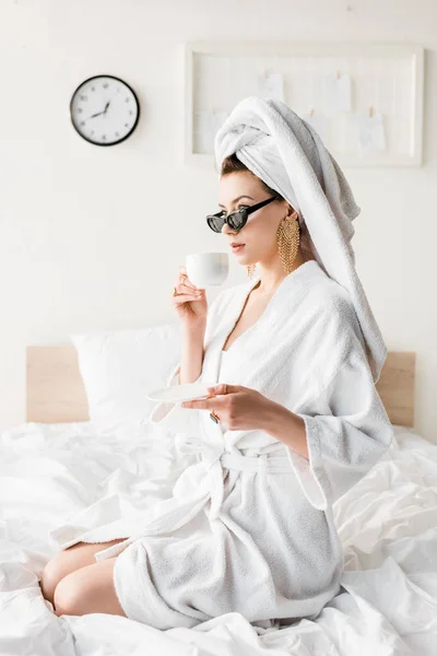 Mulher elegante em roupão de banho e óculos de sol, toalha e jóias beber café na cama — Fotografia de Stock