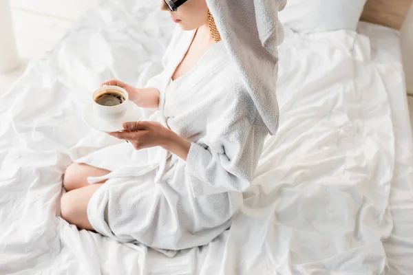 Vista recortada de la mujer con estilo en albornoz y gafas de sol, toalla y joyas beber café en la cama - foto de stock