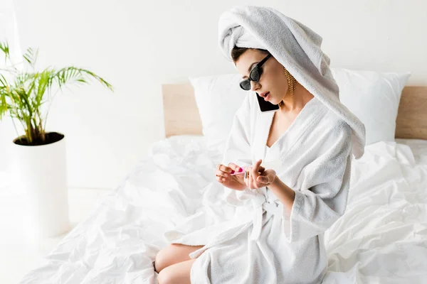 Mujer con estilo en albornoz y gafas de sol, toalla y joyas limar las uñas y hablar en el teléfono inteligente en la cama - foto de stock
