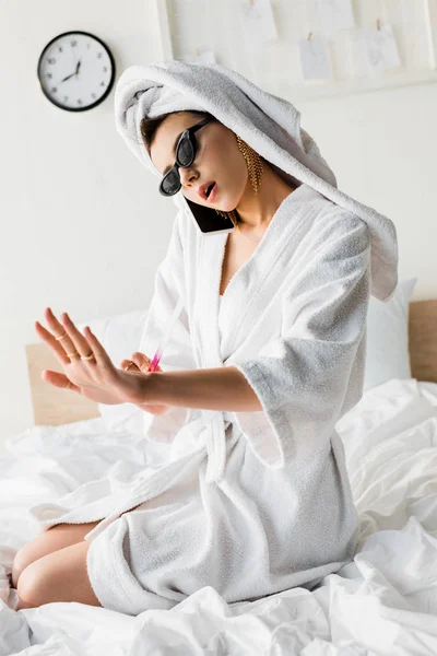 Femme élégante en peignoir et lunettes de soleil, serviette et bijoux faire manucure et parler sur smartphone au lit — Photo de stock