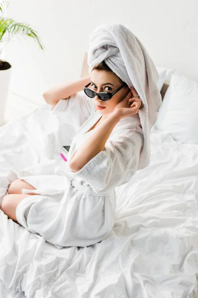 Vista aérea de la mujer con estilo en albornoz y gafas de sol, toalla y joyas sentado en la cama — Stock Photo