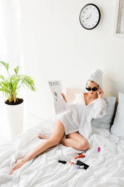 Mujer con estilo en camisa y joyas acostado en la cama con periódico de viajes cerca de esmaltes de uñas y teléfono inteligente - foto de stock