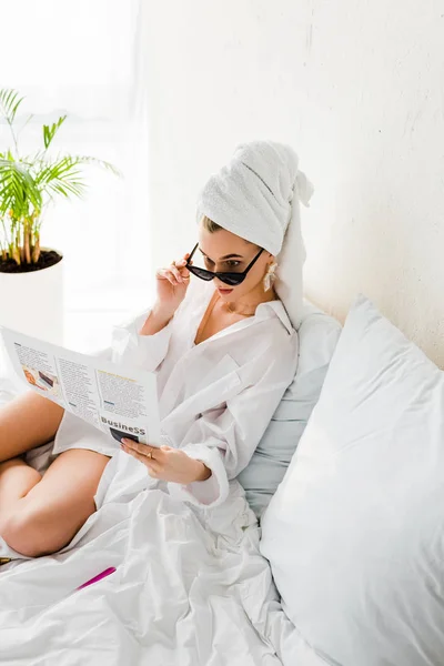 Стильна здивована жінка в сорочці, сонцезахисних окулярах та прикрасах читає газету в ліжку — стокове фото