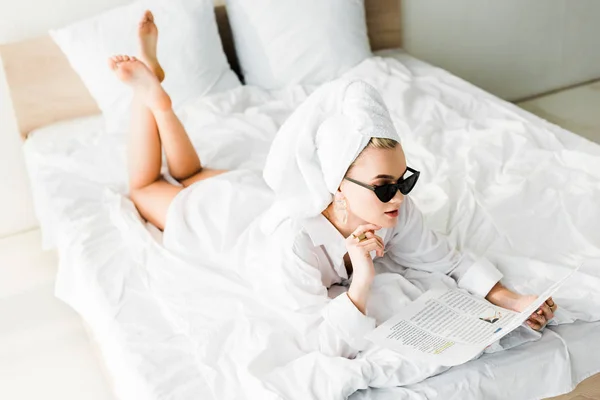 Elegante donna a piedi nudi in camicia, occhiali da sole, gioielli e con asciugamano sulla testa leggendo giornale a letto — Foto stock